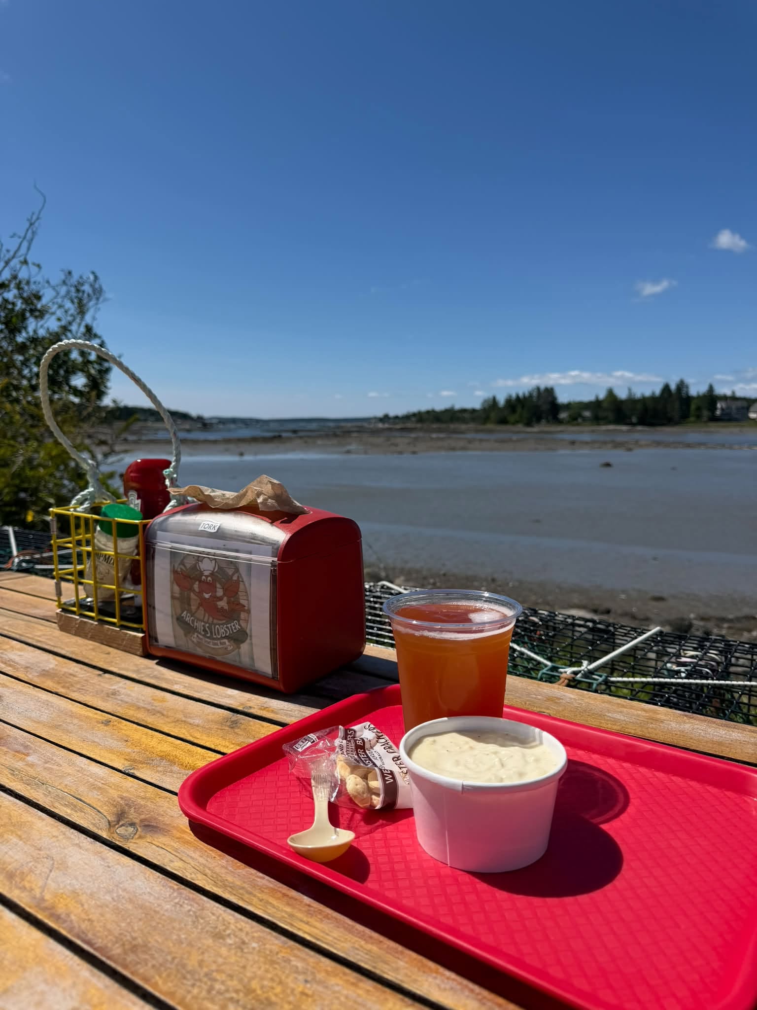 Seafood and a cold drink on a red tray at an outdoor wooden table overlooking Bass Harbor at Archie’s Lobster.
